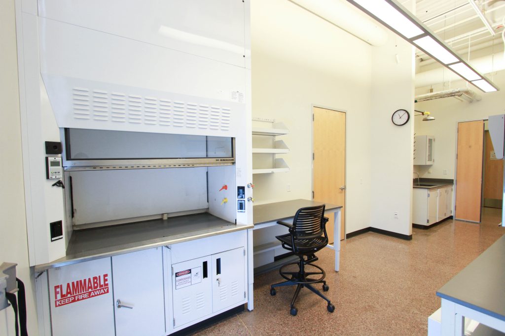 A laboratory room with a fume hood, a work desk, and a swivel chair, featuring white walls, brown tiled flooring, and ceiling fluorescent lights